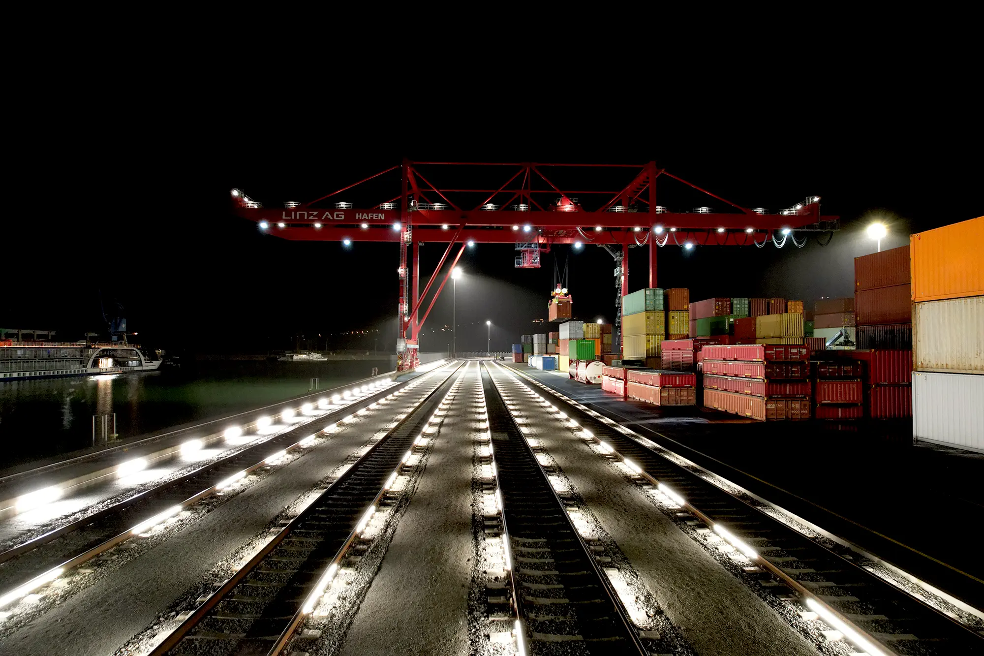 Beleuchtete Rangiergleise bei Nacht in einem Containerterminal am Hafen mit gestapelten Frachtcontainern und Containerkran. Vier Gleise Beleuchtet mit RAILUM.