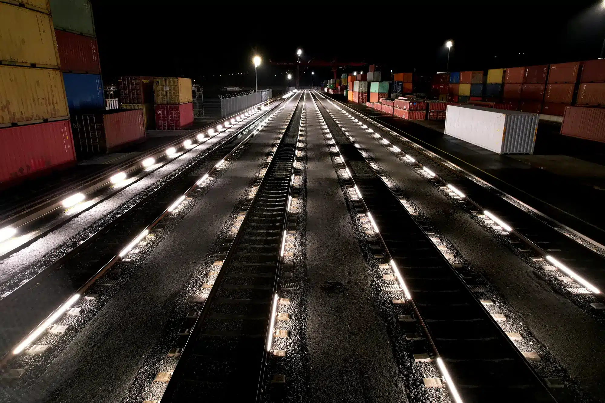 Beleuchtete Rangiergleise bei Nacht in einem Containerterminal mit gestapelten Frachtcontainern. Vier Gleise Beleuchtet mit RAILUM.