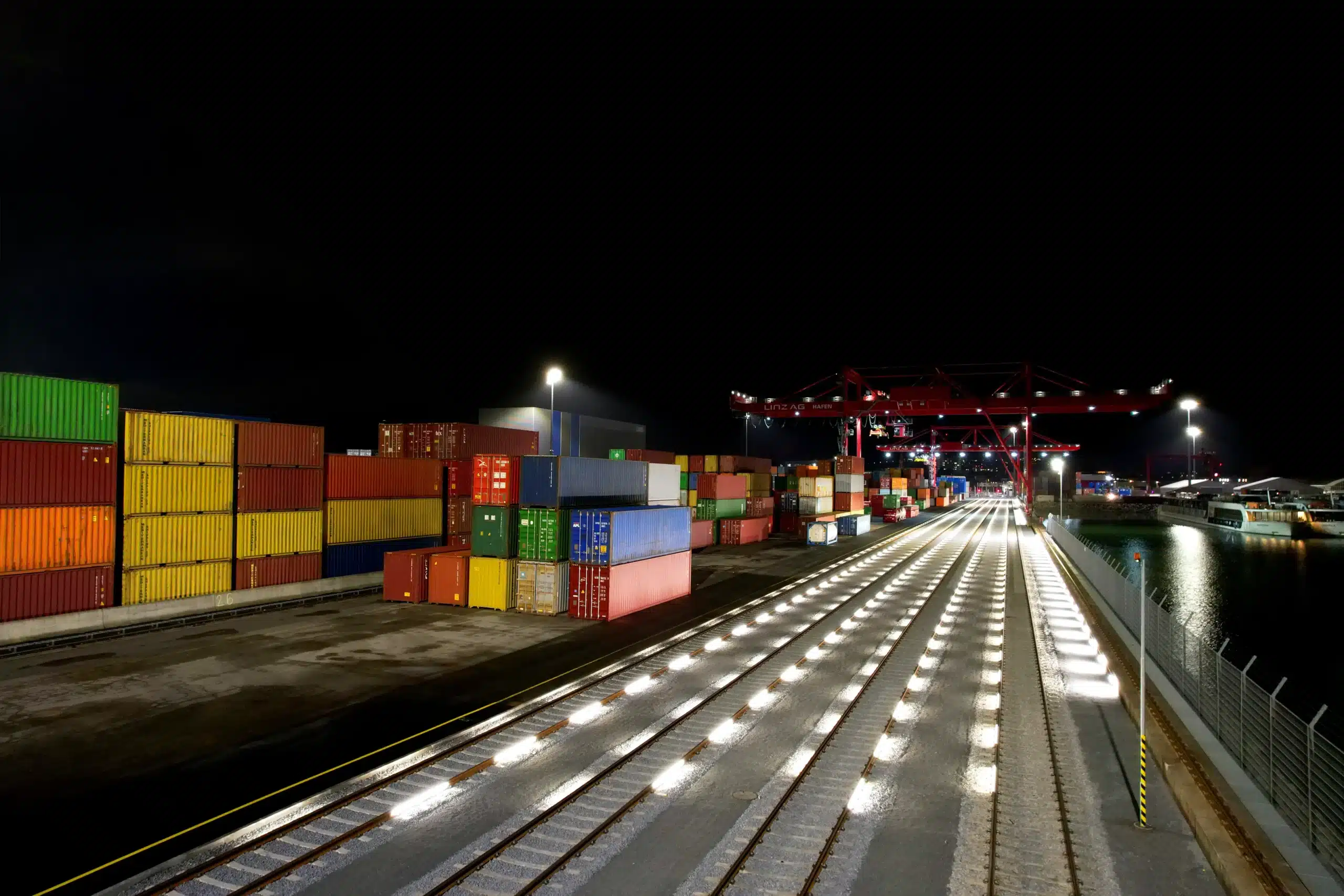 Beleuchtete Gütergleise bei Nacht im Containerterminal mit bunten Containerstapeln und Portalkran beleuchtet mit RAILUM.